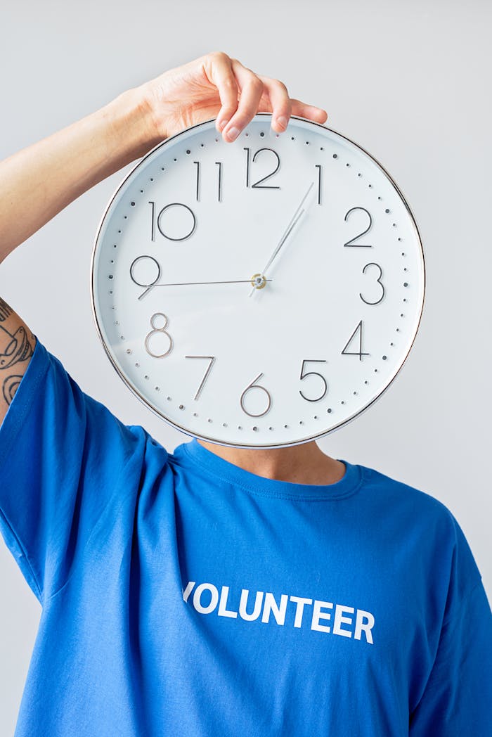 Person wearing a blue volunteer shirt holding a clock over their face against a white background.